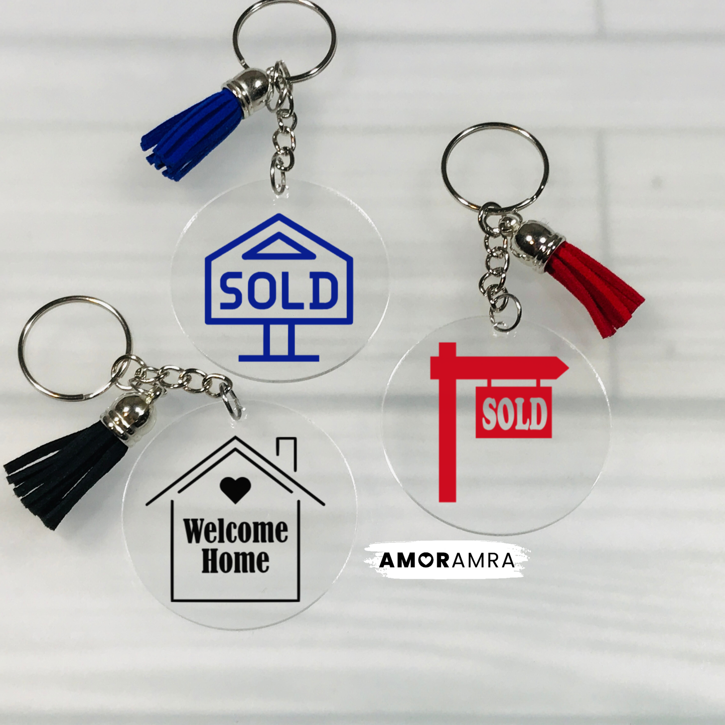 Four clear acrylic keychains with tassel charms are arranged on a white wood background. One keychain has a blue 'SOLD' house icon and blue tassel, one has a black 'Welcome Home' house icon and black tassel, one has a red 'SOLD' post and red tassel, and one is plain clear with a silver keyring and tassel. Realtor Keychain