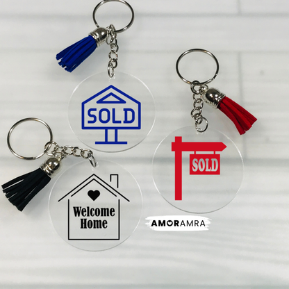 Four clear acrylic keychains with tassel charms are arranged on a white wood background. One keychain has a blue 'SOLD' house icon and blue tassel, one has a black 'Welcome Home' house icon and black tassel, one has a red 'SOLD' post and red tassel, and one is plain clear with a silver keyring and tassel. Realtor Keychain